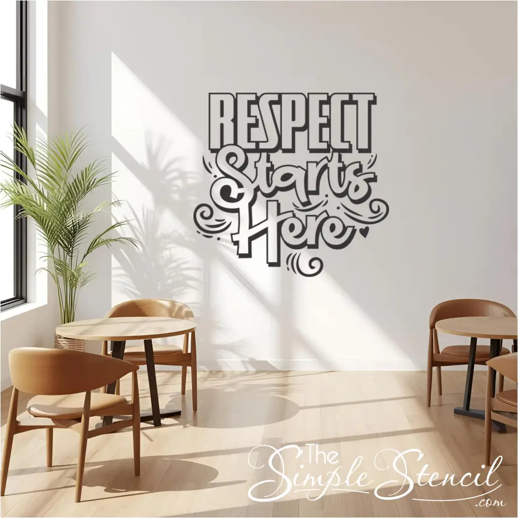 A large black "Respect Starts Here" vinyl wall decal on a white wall in a modern cafe or lounge area with wooden tables and chairs, next to a window with a green plant, illustrating respect in community spaces.