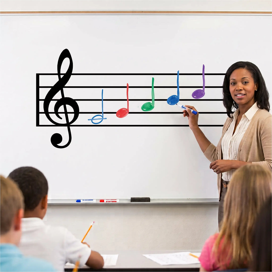 A clean, bright photograph of a large, high-quality reusable black vinyl dry-erase music staff and treble clef decal, perfectly applied to a classroom whiteboard in a modern school music room. A tray of colorful dry-erase markers below the decal illustrates its interactive function for teaching music notes and notation.