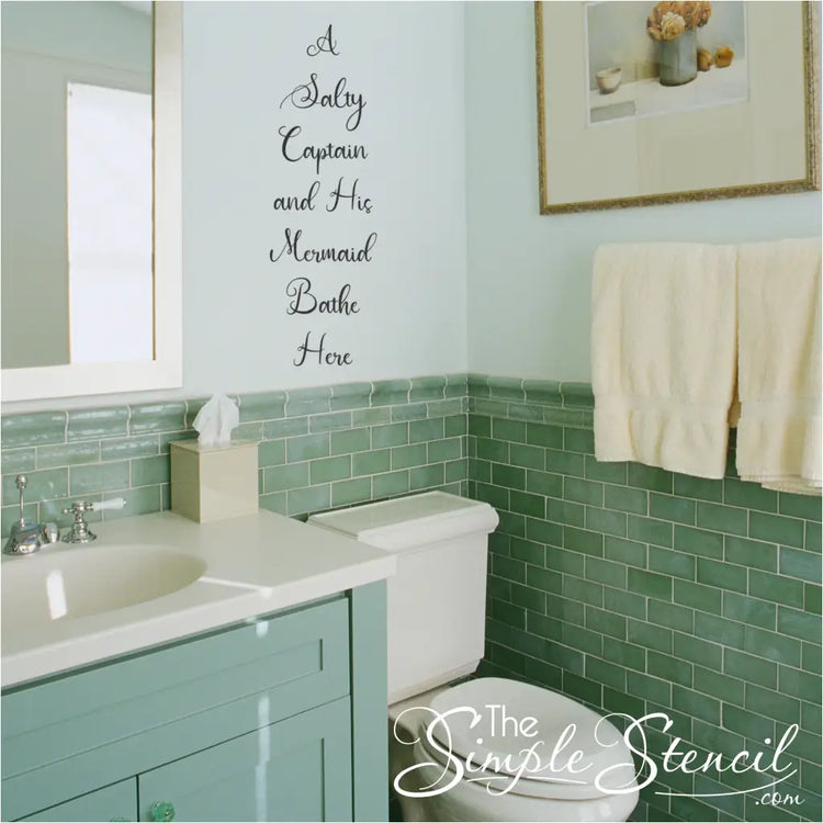 "Salty Captain & Mermaid Bathe Here" wall decal above a toilet in a beach-themed bathroom.