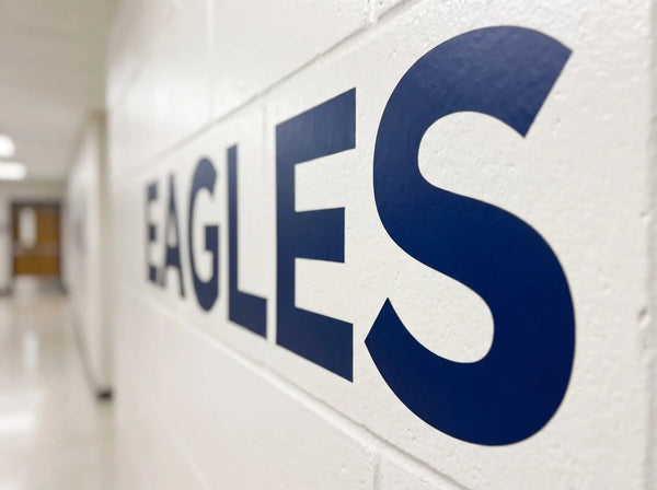 A high-resolution macro photograph capturing a detailed close-up section of a premium matte navy blue vinyl wall transfer showing the letters 'EAGLES' applied smoothly onto a painted white cinder block wall in a school hallway. The vinyl transfer flawlessly follows the subtle, natural contours and surface texture of the cinder block and paint, creating a seamless, "painted-on" appearance with zero clear plastic background.