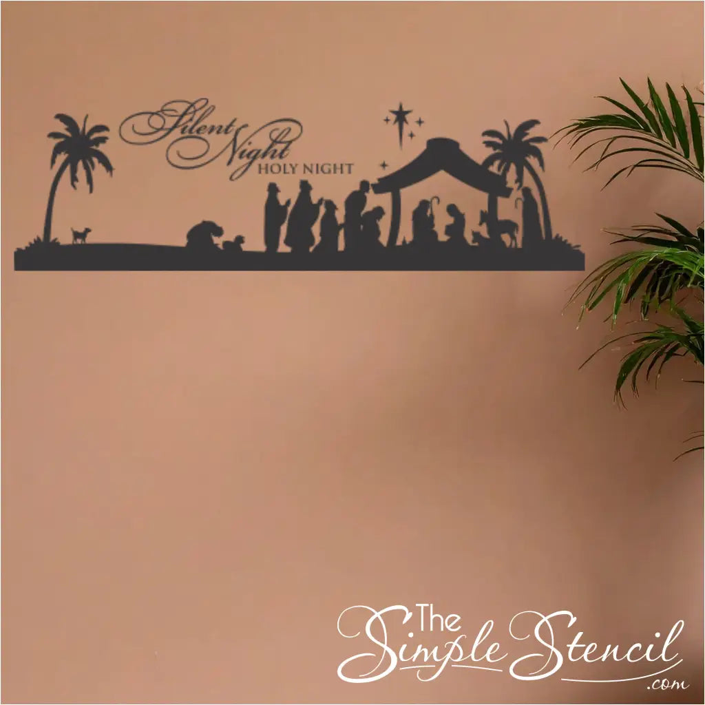 Full view of a large black nativity wall decal applied to a beige wall next to an indoor palm plant. Features the full scene and script; perfect for large Christmas decals or seasonal wall graphics in a foyer or living room.