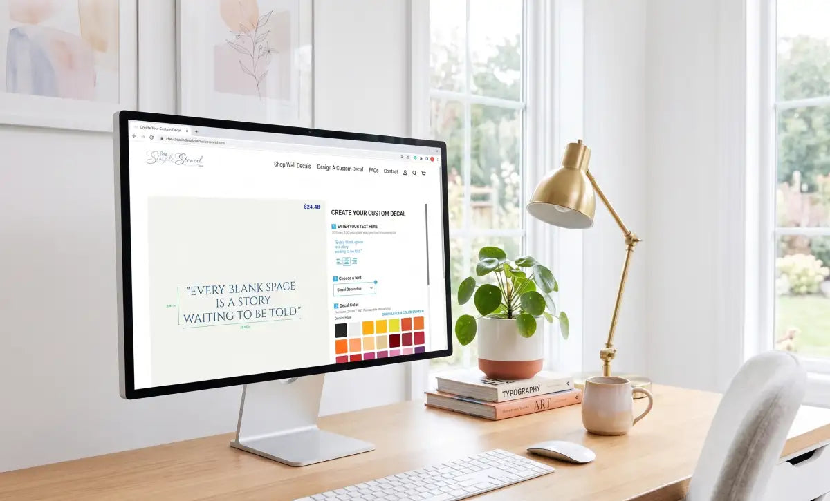 A sleek black-framed monitor displaying a website with customizable wall decals, positioned on a wooden desk beside a gold lamp and potted plant.