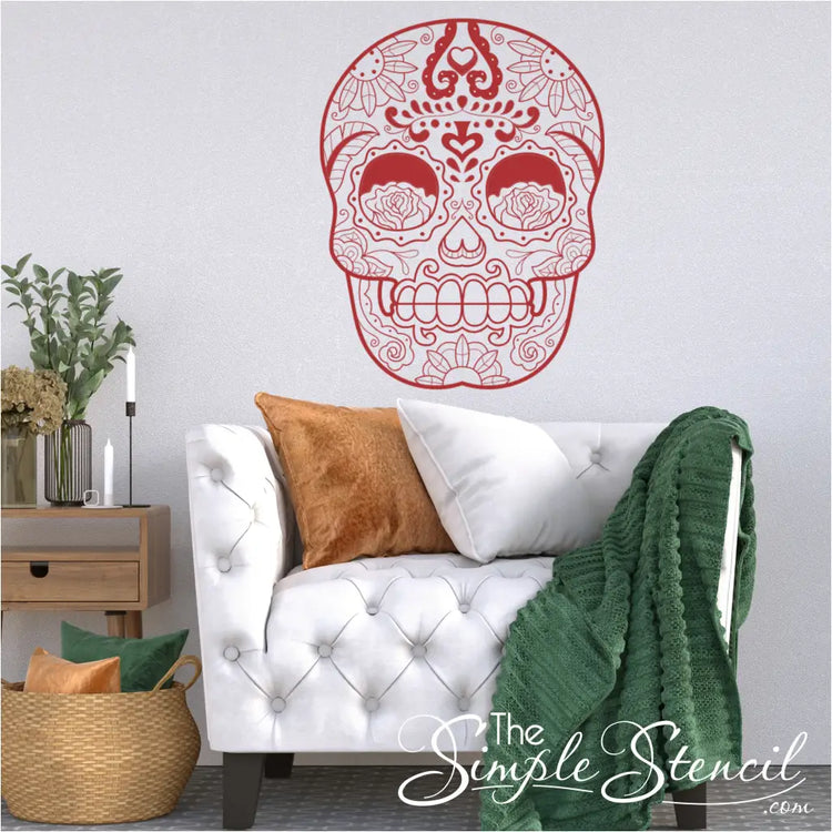  A red sugar skull vinyl wall decal placed above a white tufted sofa in a modern living room setting. Shown in 22" x 28.5" size in Merlot Red by The Simple Stencil