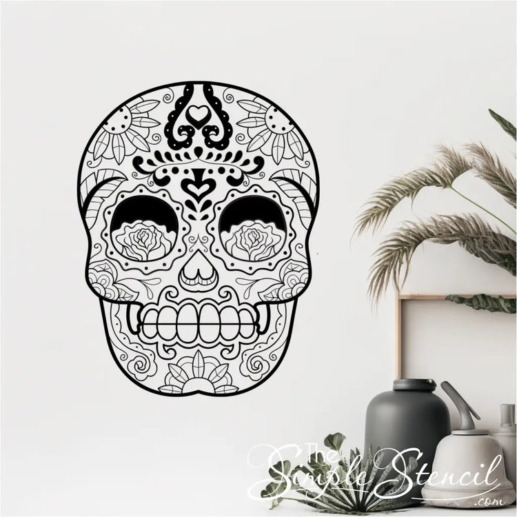A black sugar skull vinyl wall decal on a white wall, positioned next to indoor plants and modern vases. - Shown in size 22" x 28.5" size in matte black by The Simple Stencil