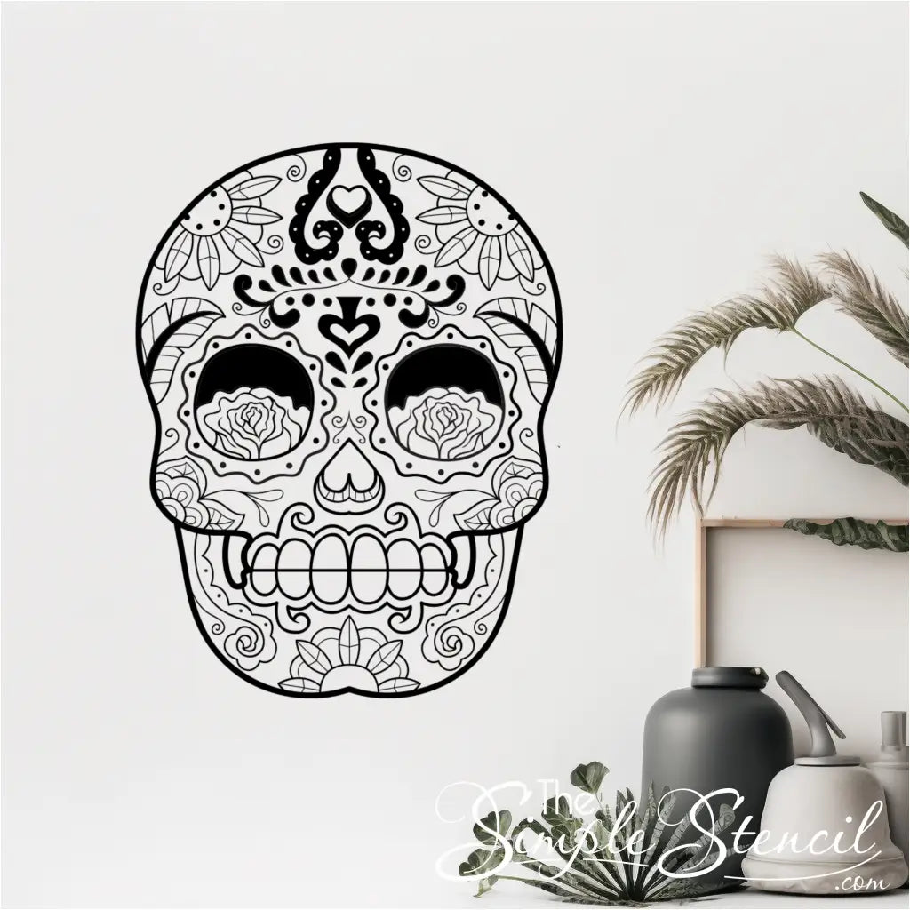 A black sugar skull vinyl wall decal on a white wall, positioned next to indoor plants and modern vases. - Shown in size 22" x 28.5" size in matte black by The Simple Stencil