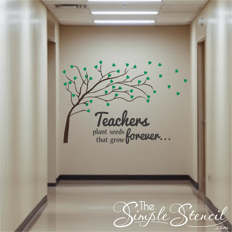 A long school hallway featuring a large wall decal of a brown tree with green heart-shaped leaves blowing in the wind. The text reads "Teachers plant seeds that grow forever..." in a mix of serif and script fonts.