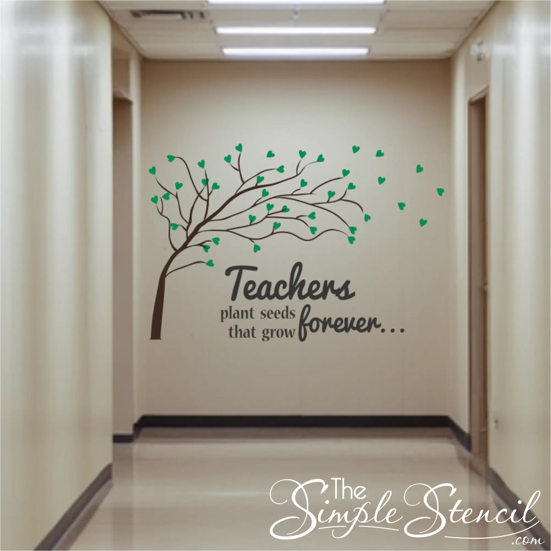 A long school hallway featuring a large wall decal of a brown tree with green heart-shaped leaves blowing in the wind. The text reads "Teachers plant seeds that grow forever..." in a mix of serif and script fonts.