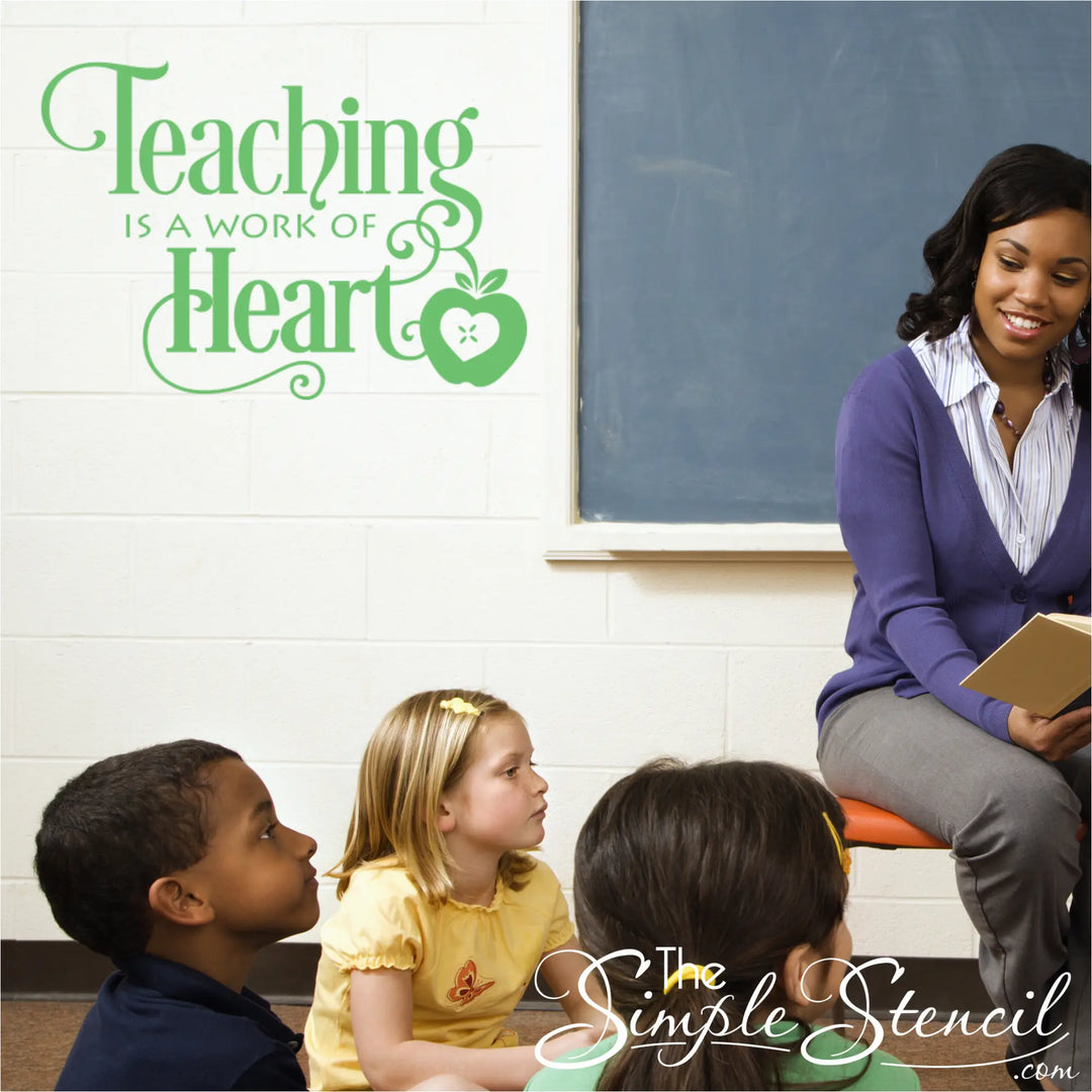 A clean, minimalist modern elementary classroom with light wood accents and sage green walls. Diverse children are seated on a calm grey rug listening to a teacher read. A large sage green vinyl wall stencil reading 'Teaching is a Work of Heart' is applied to the main white block wall above them.