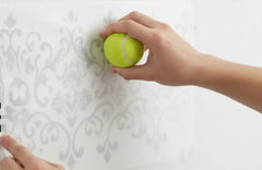 A hand pressing a vinyl decal through its transfer tape with a tennis ball to ensure it adheres to a textured wall.