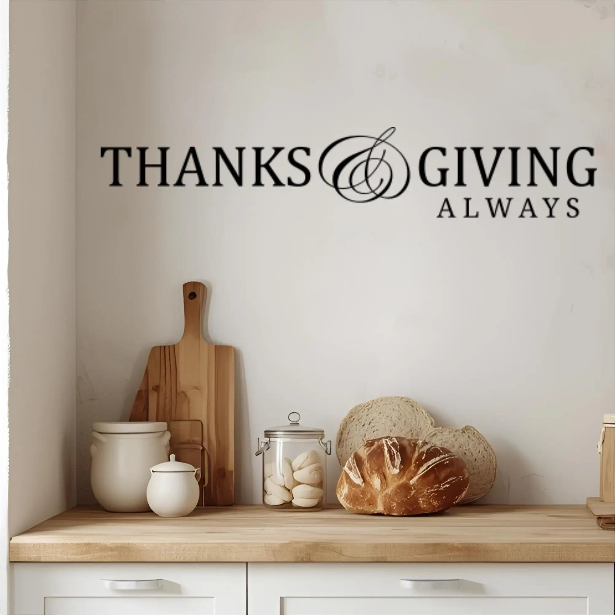 The black 'Thanks and Giving Always' wall decal is displayed on a kitchen wall above a wooden countertop with bread and white ceramic jars, creating a modern farmhouse look.