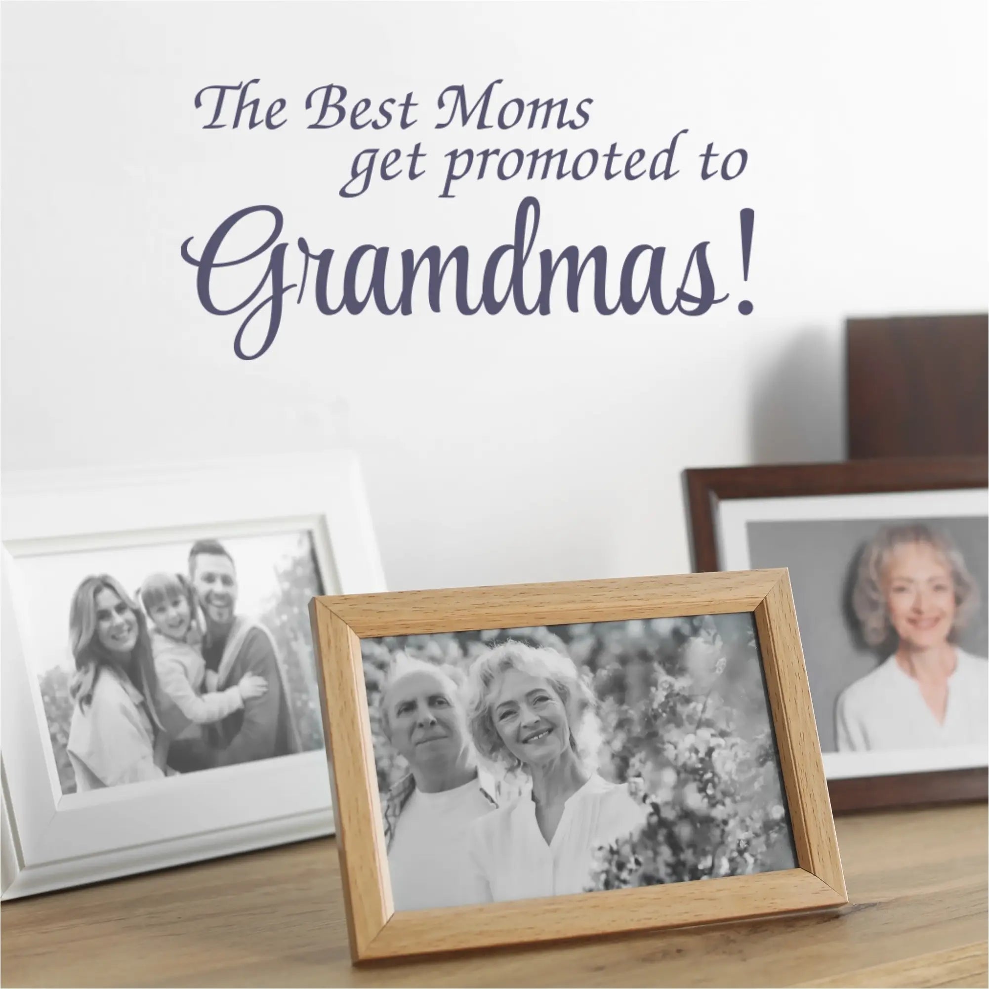 "The Best Moms Get Promoted to Grandmas!" wall decal in navy blue vinyl applied to a kitchen wall, with a vase of flowers and a framed family photo nearby.