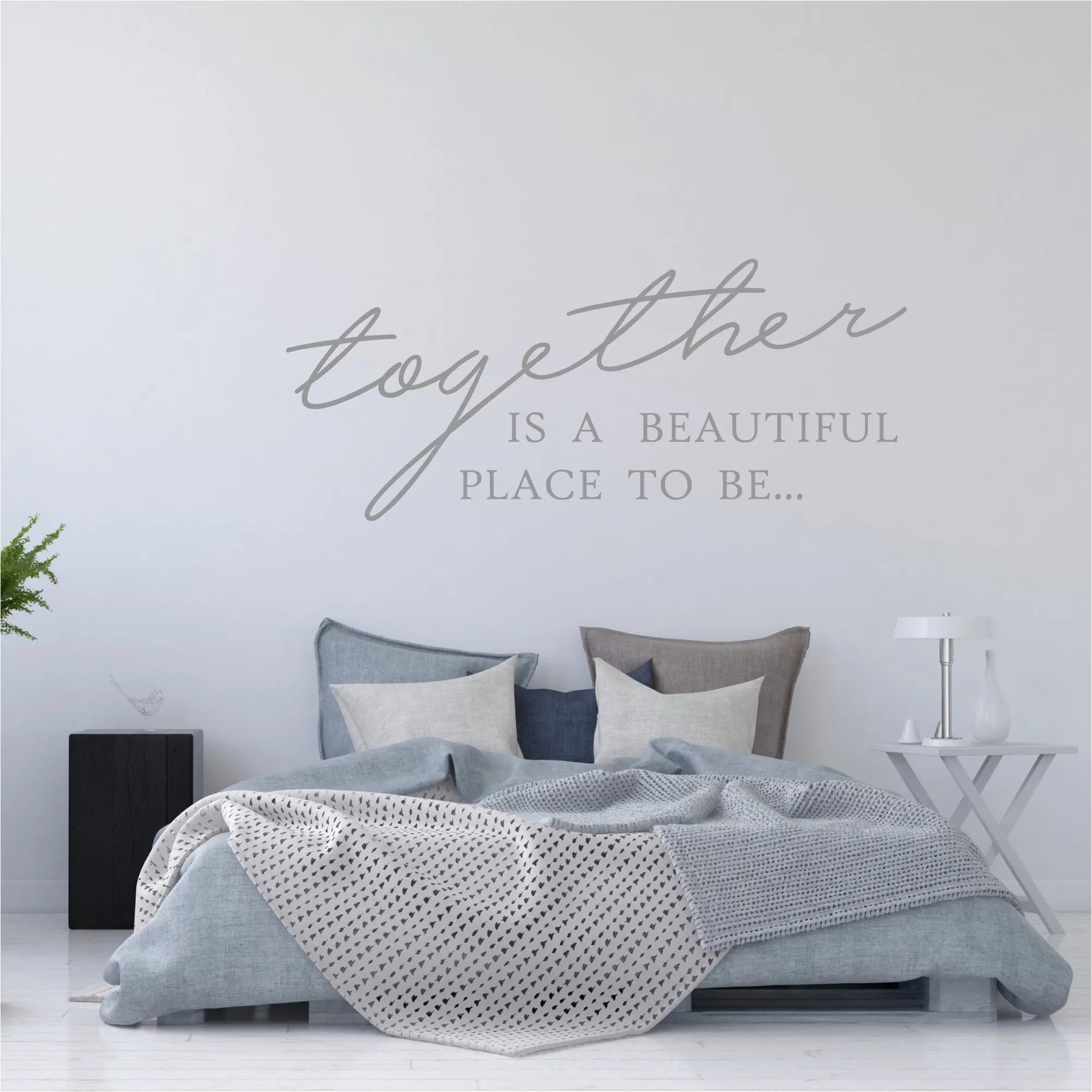 Minimalist bedroom with light blue bedding and a grey "Together is a beautiful place to be" wall decal centered on a clean white wall.