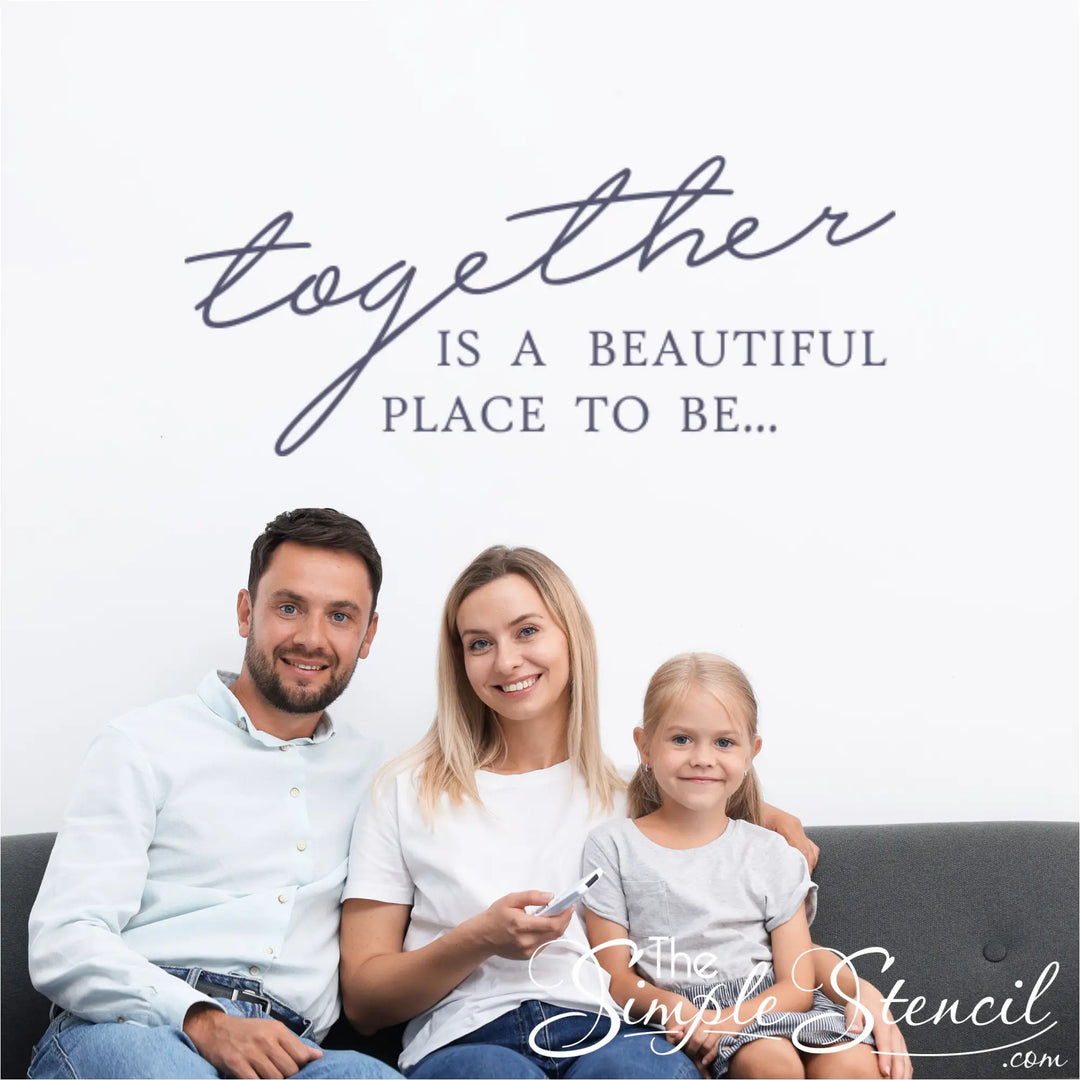 Happy family of three sitting on a grey sofa beneath a large navy blue wall decal that reads "Together is a beautiful place to be," showcasing family room decor.