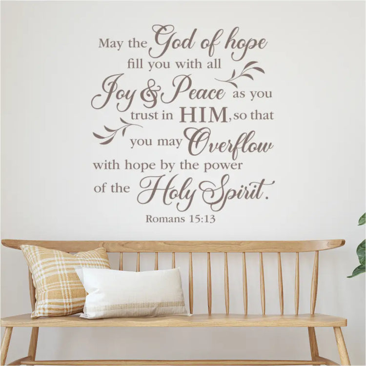 Large, elegant black vinyl wall decal of Romans 15:13 "May the God of hope..." on a white wall above a wooden bench in a modern entryway.