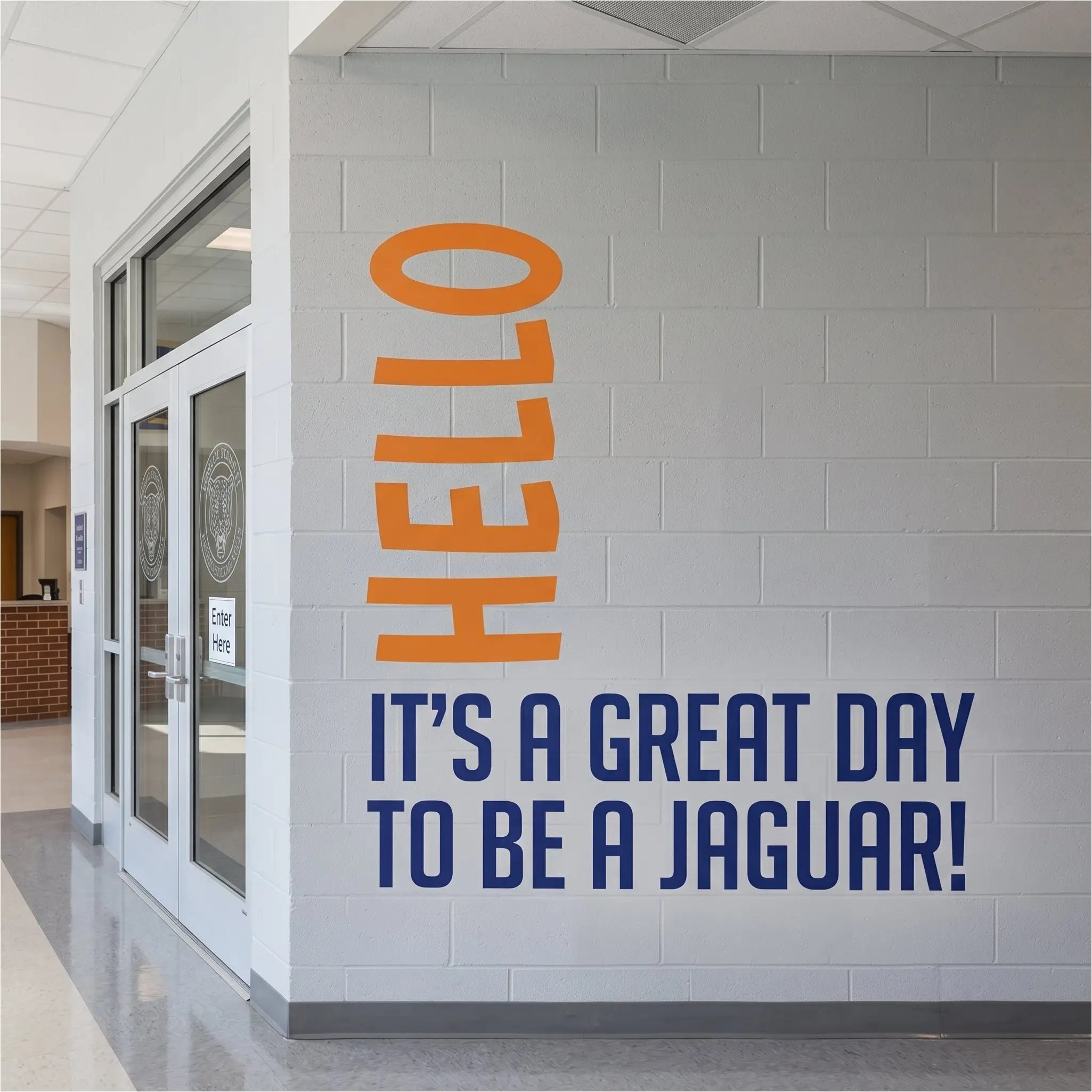 A school hallway wall with a white brick pattern, featuring large orange vertical 'HELLO' lettering and navy blue text that reads 'IT'S A GREAT DAY TO BE A JAGUAR!'.