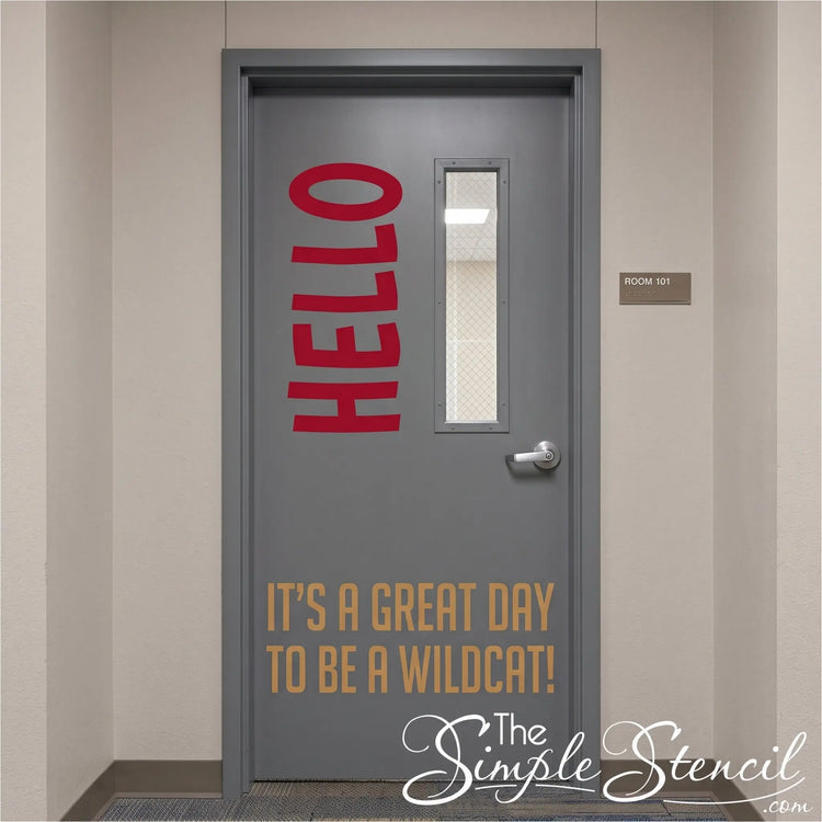 A gray office-style door with a narrow vertical window, silver handle, and printed text reading 'HELLO' in red vertically on the upper left and 'IT'S A GREAT DAY TO BE A WILDCAT!' in tan near the bottom. 