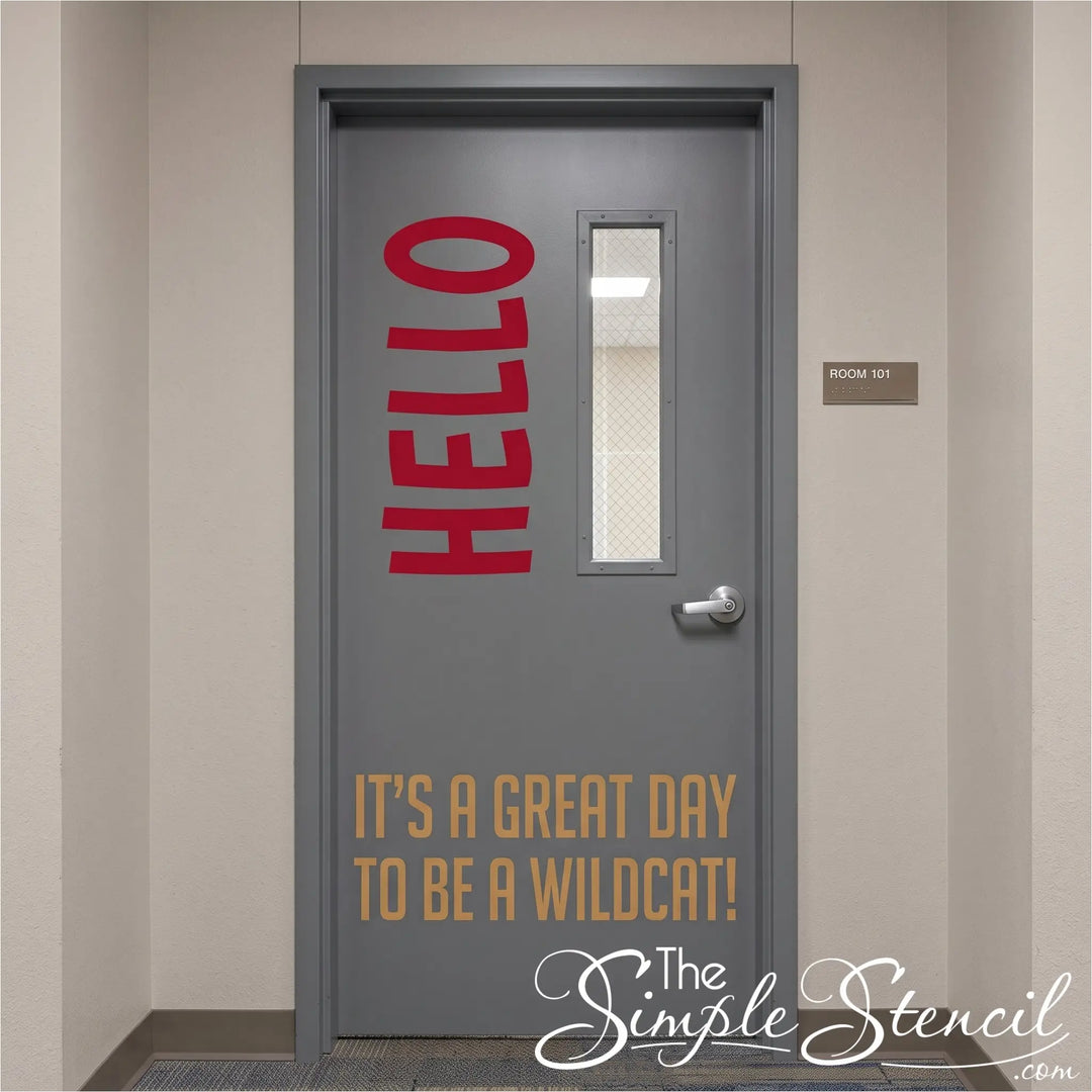 A gray office-style door with a narrow vertical window, silver handle, and printed text reading 'HELLO' in red vertically on the upper left and 'IT'S A GREAT DAY TO BE A WILDCAT!' in tan near the bottom. 
