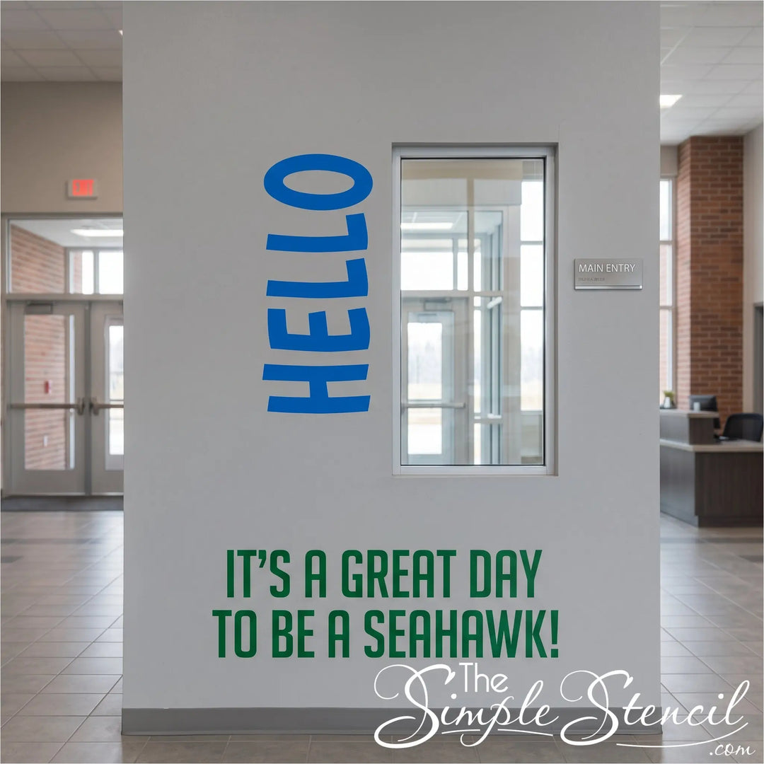 A white interior wall in a modern building lobby features large blue vertical 'HELLO' lettering and green text reading 'IT'S A GREAT DAY TO BE A SEAHAWK!' near the bottom.