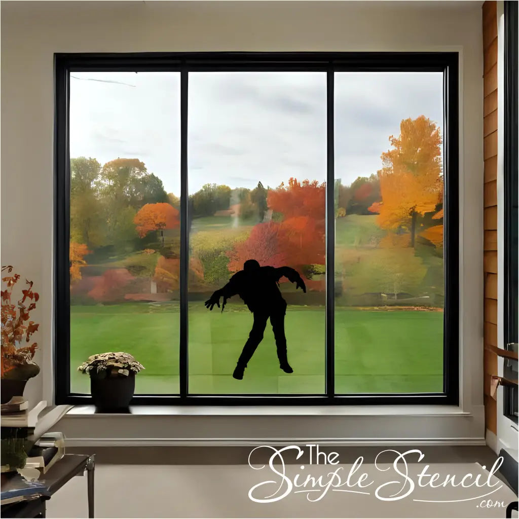 A black walking zombie silhouette vinyl decal applied to a glass patio door, with a cozy indoor autumn setting and a view of fall trees.