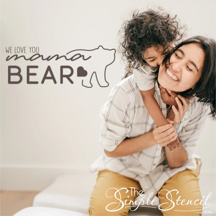 "We Love You Mama Bear" wall decal in a warm brown vinyl applied to a living room wall, with a cozy armchair and throw pillows nearby a mom and son playing. By The Simple Stencil
