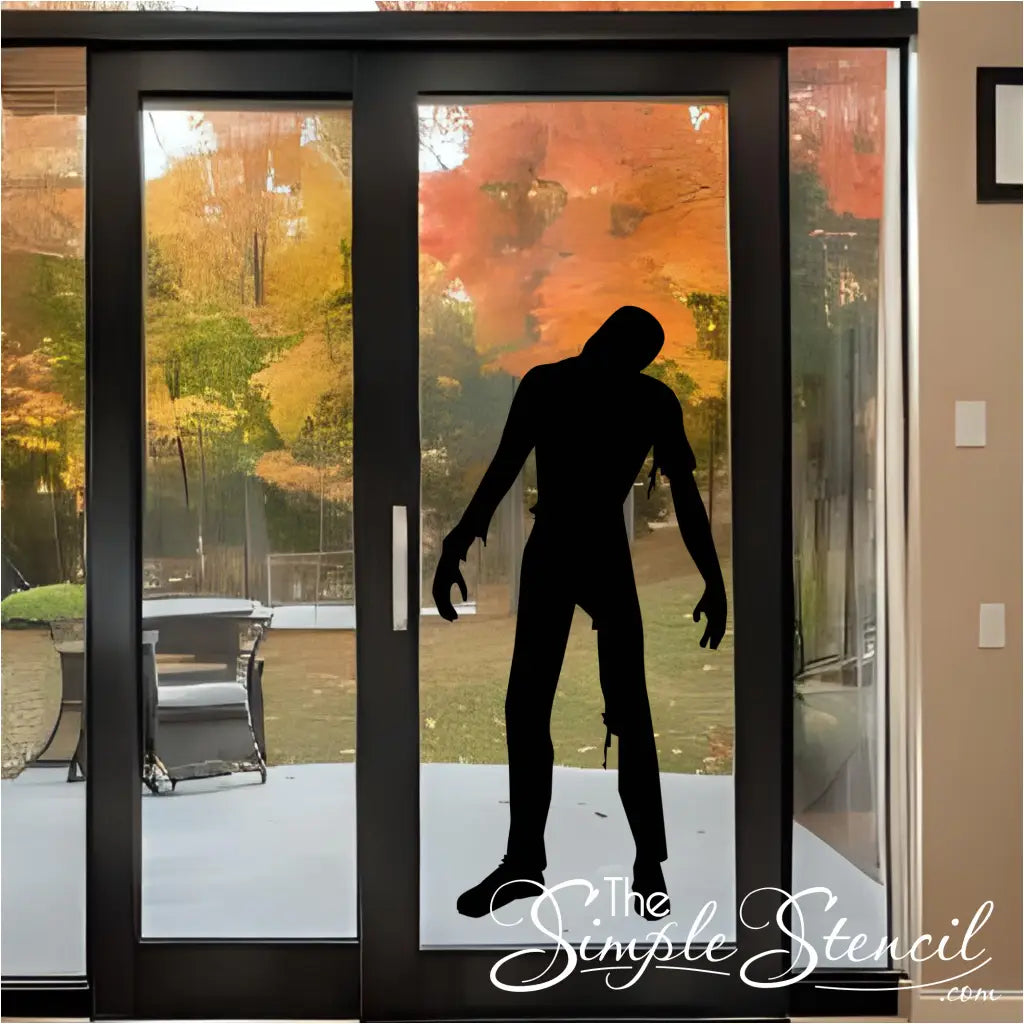 A black zombie silhouette vinyl decal on a modern black-framed patio door, with an outdoor patio and fall trees in the background, creating a scary Halloween scene.