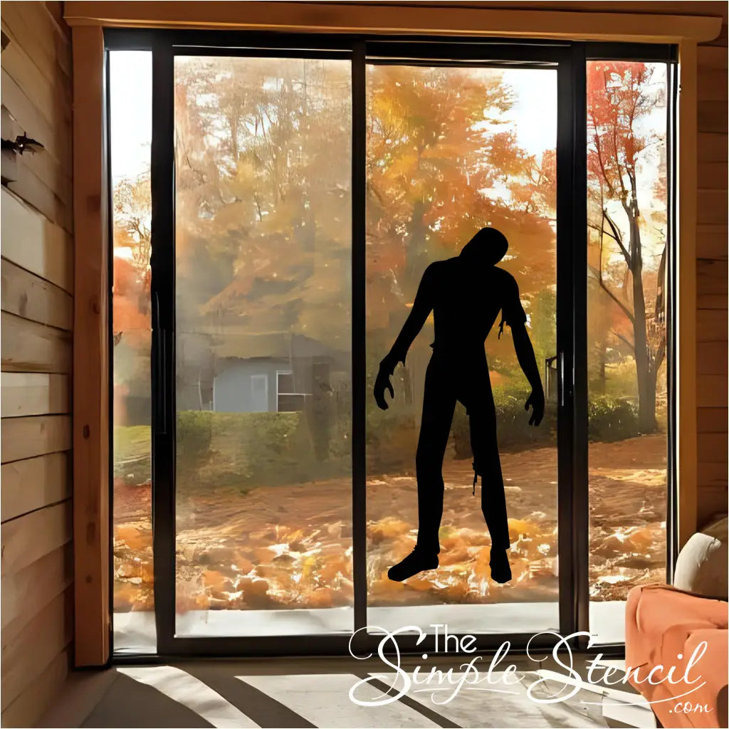 A black zombie silhouette vinyl decal applied to a glass patio door, overlooking an autumn landscape with fall foliage, adding a spooky touch to home decor. By The Simple Stencil